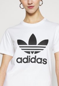 Balta kokvilnas T-krekls ar izteiktu melnu Adidas logo, trīs svītrām un trifeles dizainu priekšpusē. Apaļš kakls un īsas piedurknes.