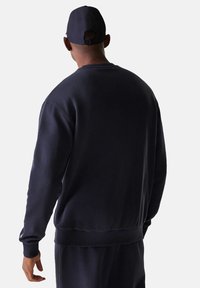 Hombre con sudadera azul marino, pantalones a juego y gorra de béisbol azul marino de espaldas contra un fondo claro y sencillo.