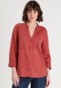 Chemise à manches longues de couleur rouille, avec un devant boutonné, un col à encoche, et deux poches poitrine. Fabriquée en tissu léger et texturé.