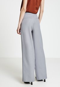 Pantaloni grigio chiaro a gamba larga con finitura testurizzata, caratterizzati da una vita alta e due tasche posteriori. Indossati con una maglia marrone.