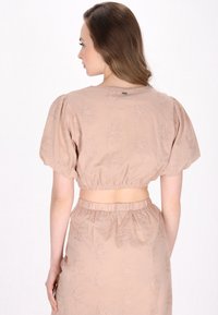 Blush-farvet cropped top med puffærmer, blomsterbroderi og elastisk bånd i taljen. Synlig lukning ved nakkedetaljen.