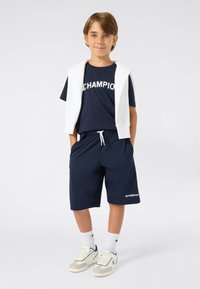 Marine Champion T-shirt met witte letters, bijpassende marine korte broek met zijzakken, witte trui over de schouders gedrapeerd en witte sneakers.
