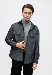 Fiatal férfi sötétszürke gombolós felsőruházatot visel világosszürke garbó pulóver és fekete nadrág felett, egyszínű háttér előtt áll.