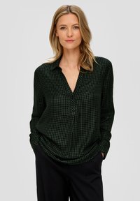 Ciemnozielona bluzka w wzór houndstooth, z długimi rękawami, kołnierzykiem i luźnym krojem, w zestawieniu z ciemnymi spodniami.