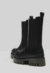 Stivaletti neri in pelle con pannelli laterali elasticizzati e una suola platform grigia alta con scanalature profonde. Texture liscia, design minimale.