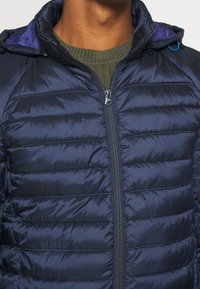 Marineblå dunjakke med syede paneler, lynlås og hætte. Har et tekstureret design og glat ydre stof.