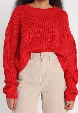 Maglione rosso lavorato a maglia con un design morbido e oversize, caratterizzato da maniche lunghe e un motivo testurizzato, abbinato a jeans beige a vita alta.