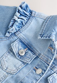 Giacca di jeans azzurro chiaro con bottoni argento, tasca sul petto, dettaglio arricciato vicino alla spalla e collo piegato su uno sfondo chiaro.
