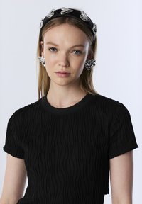 Zwart textuur top met een golfpatroon, gecombineerd met zilveren hartvormige oorbellen en een zwarte haarband met metallic accenten.