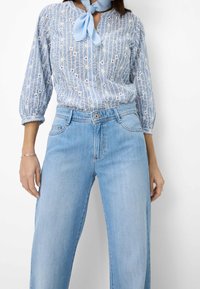 Blusa azul claro con bordado geométrico blanco y mangas de 3/4, combinada con jeans de tiro alto azul claro con diseño clásico de cinco bolsillos.