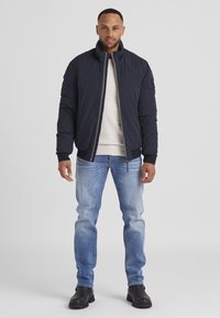 Marineblauer Pufferjacke mit hohem Kragen und gerippten Bündchen, kombiniert mit hellblauen Jeans und schwarzen Knöchelstiefeln. Einfacher Reißverschluss und elegantes Design.