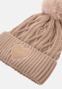 Beige gestrickte Mütze mit Kabelmuster, umgeschlagenem geripptem Bündchen und einem braunen Herz-Patch, das "GUESS" in erhabenem Schriftzug und Verzierungen zeigt.