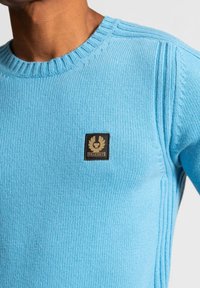 Man som bär en ljusblå stickad tröja med ribbad rundhals och svart fyrkantig Belstaff-logotyp på bröstet.