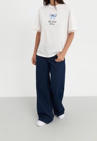 Osoba ubrana w luźny biały T-shirt z niebieską kokardą i napisem "Powinieneś ze mną chodzić", szerokie ciemnoniebieskie dżinsy i białe trampki.