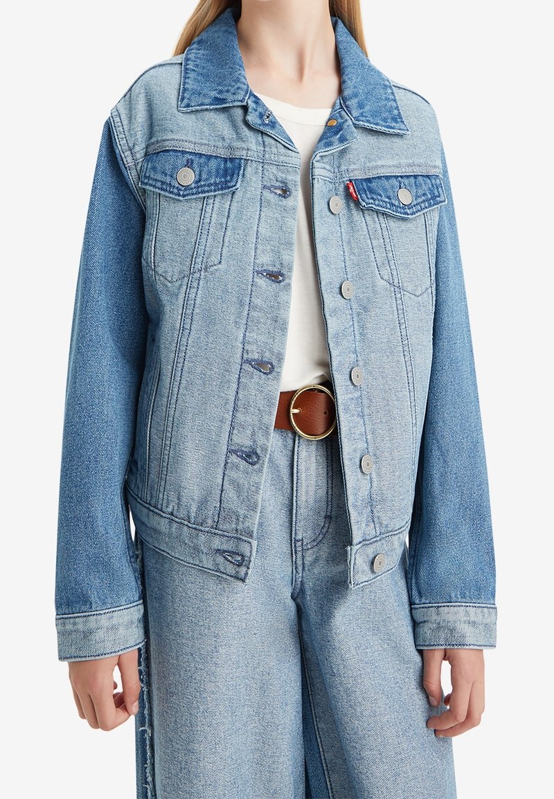 Veste en denim bicolore bleu clair avec boutons, poches poitrine à rabat, portée sur un top blanc et associée à un pantalon en denim assorti et une ceinture marron.