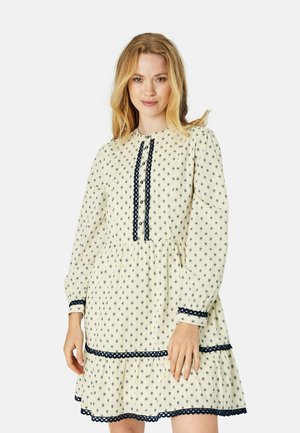 Floral-patterned cream dress with navy accents, featuring a high neckline, button placket, and tiered ruffle hemline. Long sleeves.
