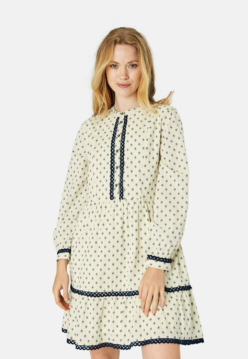 Cremefarbenes Kleid mit floralen Mustern und marineblauen Akzenten, ausgestattet mit einem hohen Kragen, einer Knopfleiste und einem gestuften Rüschensaum. Lange Ärmel.