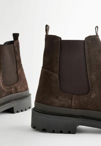 Botine maro din suede cu părți elastice, tab-uri întărite la călcâi și talpă din cauciuc texturat negru. Prezintă un design solid și clasic.