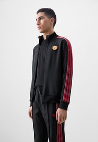 Black athletic jacket with red striped accents, zip closure, standing collar, and embroidered logo. Paired with matching pants featuring logo.