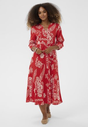 Ženska s kodrastimi lasmi, oblečena v rdečo midi obleko s cvetličnim vzorcem in rokavi do komolcev, ter bež tkanimi balerinkami, stoji na belem ozadju.