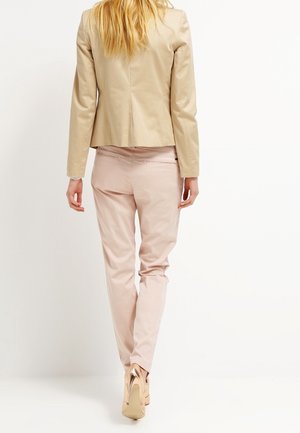 Mujer caminando de espaldas con chaqueta beige, pantalones rosa claro y zapatos de tacón dorados sobre un fondo blanco liso.