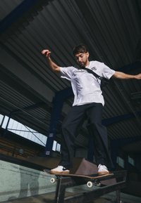 Skateboarder iført en hvid Nike SB t-shirt og sorte bukser udfører et trick på et rækværk i en indendørs skatepark med metal- og træoverflader.