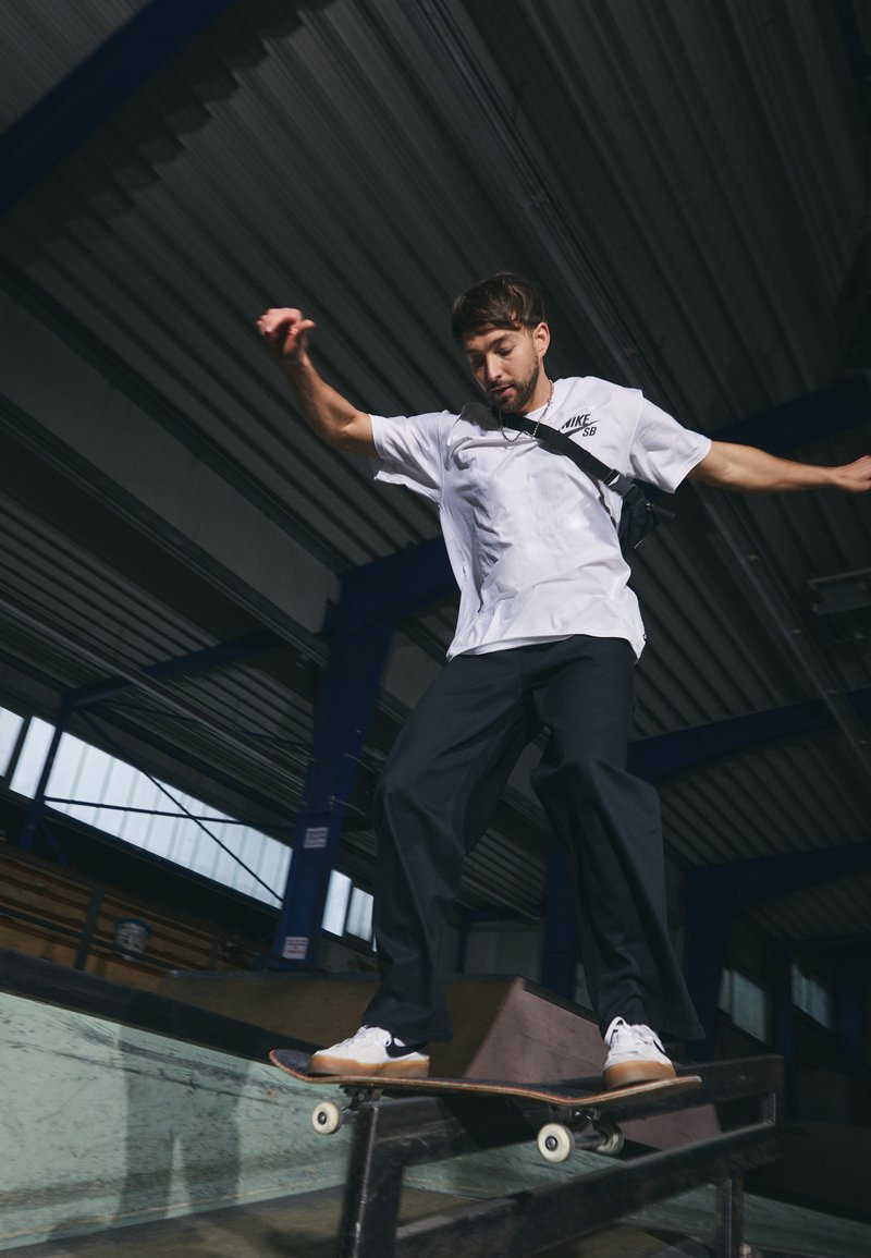 Skater en una camiseta blanca de Nike SB y pantalones negros realiza un truco en una barandilla en un skate park interior con superficies de metal y madera.
