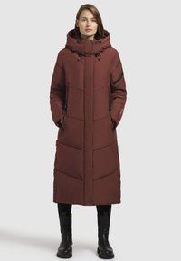 Geacă puffer lungă, roșie cărămizie, cu glugă și buzunare cu fermoar. Dispune de cusături în formă de V și nasturi cu capsa pe lateral. Pur tată peste pantaloni negri și cizme.
