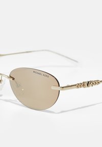 Zelta metāla saulesbrilles ar ovālām brūnām lēcām. Iespējas ietver zīmola nosaukuma "MICHAEL KORS" drukāšanu uz lēcas un rakstainu tempļa dekoru.