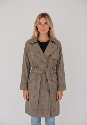 Mujer con cabello rubio largo que lleva un abrigo estilo trench a cuadros con cinturón sobre una camiseta negra y jeans azul claro, de pie contra un fondo liso.