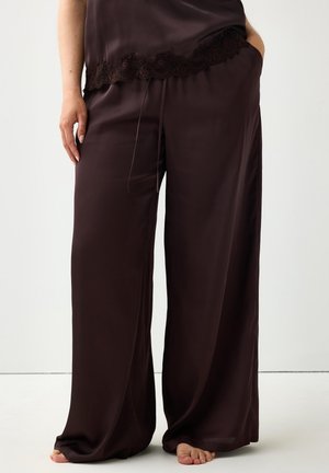Donna che indossa pantaloni larghi in satin marrone scuro con coulisse in vita e top con bordo in pizzo, in piedi a piedi nudi su uno sfondo semplice.