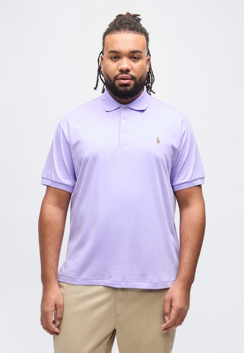 Άνδρας με πλεγμένα μαλλιά και γένια, φορώντας ανοιχτό μωβ πόλο πουκάμισο και μπεζ παντελόνι, στέκεται μπροστά σε απλό λευκό φόντο.