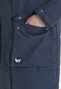 Mörkblå vattentålig jacka med tryckknappsstängning, utrustad med en framficka och en logotypflik märkt "WHISTLER" i framträdande läge.