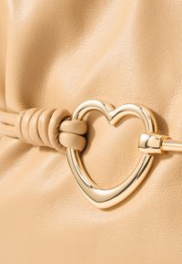 Μπεζ δερμάτινη τσάντα χειρός με χρυσό μπουτόν σε σχήμα καρδιάς και λεπτομέρεια από δερμάτινο κόμπο στη λαβή, τονίζοντας την υφή και την ομαλή επιφάνεια.