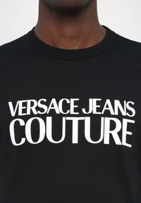 Juodas medvilnės marškinėlis su dideliu baltu spaudiniu, kuris skaito "VERSACE JEANS COUTURE" per krūtinę. Paprasta apvalios iškirptės forma.
