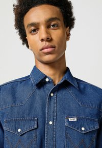 Camicia in denim blu con design del giogo a punta, bottoni a pressione bianchi e due tasche sul petto. Etichetta del logo Wrangler sulla tasca sinistra.