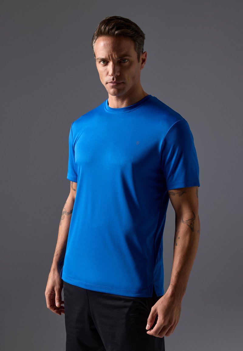 Sinine sporditshirt ümmarguse kaeluse, lühikeste varrukate ja tekstuurse materjaliga; koos mustade lühikeste pükstega. Minimimaalne bränding on nähtav.