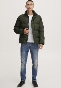 Green puffer jacket with a stand-up collar, button closure, and a small logo patch, paired with light blue jeans and black sneakers.