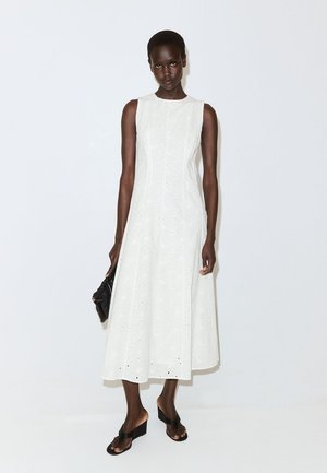 Donna che indossa un vestito midi bianco senza maniche con dettagli a occhiello, tiene una piccola clutch nera e indossa sandali con zeppa neri.