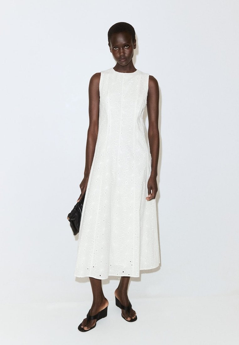 Femme portant une robe midi blanche sans manches avec des détails en broderie anglaise, tenant une petite pochette noire et portant des sandales compensées noires.