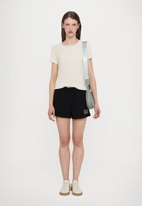 Krämfärgad t-shirt, svarta shorts med justerbart snöre, guldbroderad detalj, beigefärgade sneakers och en ljusgrön axelväska med rem.