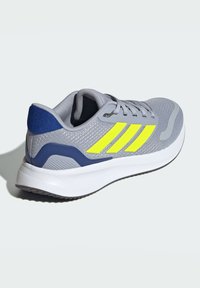 Zapatilla deportiva gris con diseño de tres rayas amarillas, parte superior de malla texturizada, tapa de talón azul y media suela blanca.