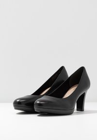 Pár černých kožených dámských lodiček na středním podpatku s kulatou špičkou na bílé ploše na pozadí s jednobarevným světlým šedým pozadím.