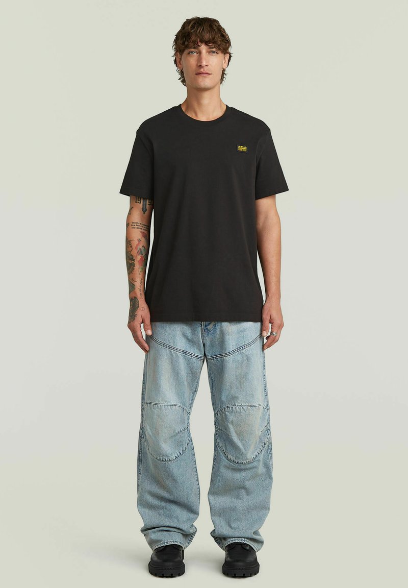 Giovane uomo con capelli ricci che indossa una T-shirt nera e jeans azzurri larghi, in piedi con le braccia rilassate ai lati.