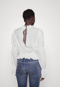 Blouse blanche avec un col volanté et une ouverture dans le dos, dotée d'une fermeture à nouer. Manches longues bouffantes et taille smockée. Associée à un jean bleu.