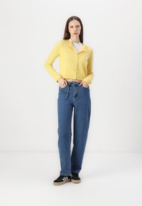 Cardigan amarelo de malha com fecho em botão, combinado com jeans azuis de cintura alta e ténis pretos com riscas brancas. Fundo simples.