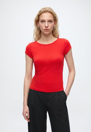 Femme blonde portant un haut rouge ajusté à manches courtes et un pantalon noir, debout avec une main dans la poche sur un fond blanc uni.