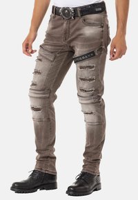 Graue, beschädigte Jeans mit Ripped-Details,Slim-Fit und strukturierten Patches. Kombiniert mit einem schwarzen Gürtel und schwarzen Schnürboots.
