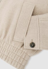 Beige strukturierte Jacke mit Kragen, mit einem schwarzen Knopf und elastischem Bündchen. Der Stoff hat ein gewebtes Muster und einen gerafften Saum.