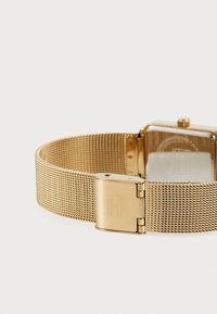 Montre-bracelet en maille dorée avec un cadran carré et une fermeture à clip. Le bracelet présente un motif texturé, complétant la finition brillante de la montre.